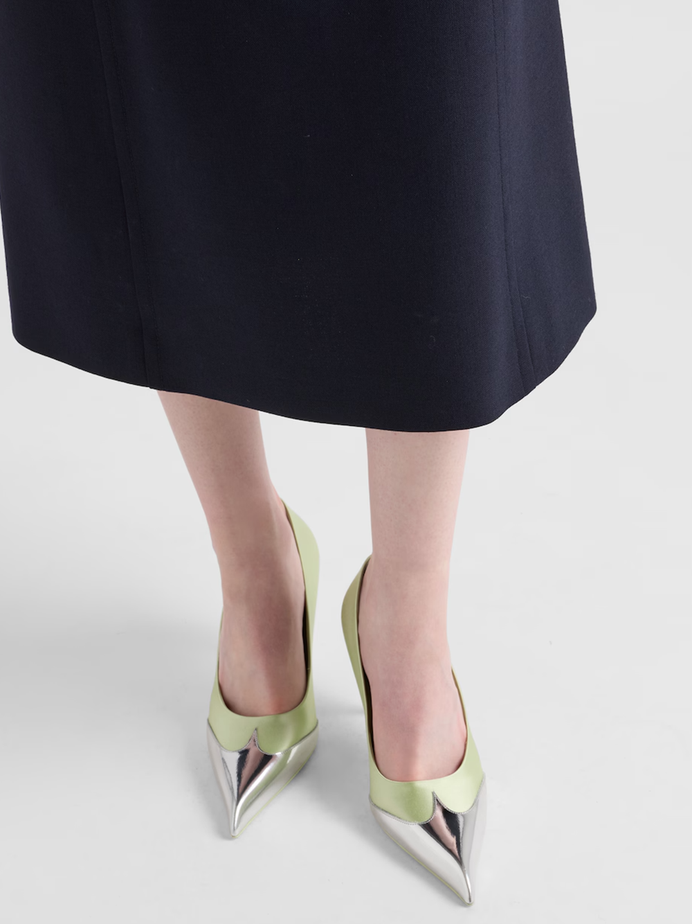 Green high-heeled shoes with metallic tips worn by a person in a black skirt on a light gray background